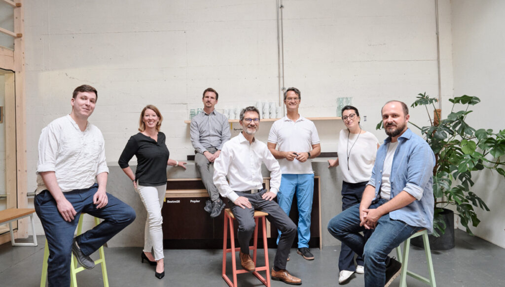 Das Team von Valean Solutions aus München posiert mit sieben Personen in einem modernen Büro mit industriellem Design in München. Sie stehen und sitzen entspannt auf Barhockern und wirken professionell und freundlich.