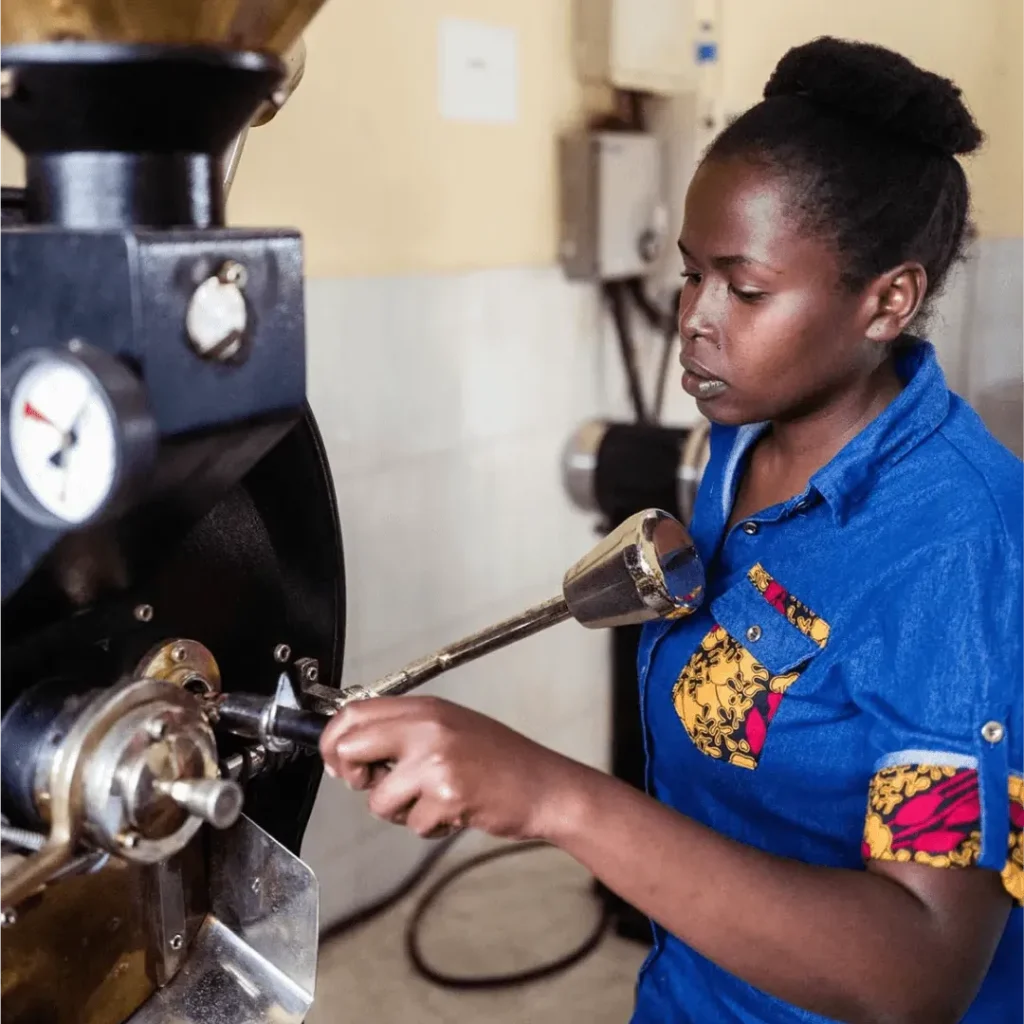 Eine Frau in einer blauen Bluse mit bunten Mustern überprüft den Röstgrad der Kaffeebohnen an einer Kaffeeröstmaschine.